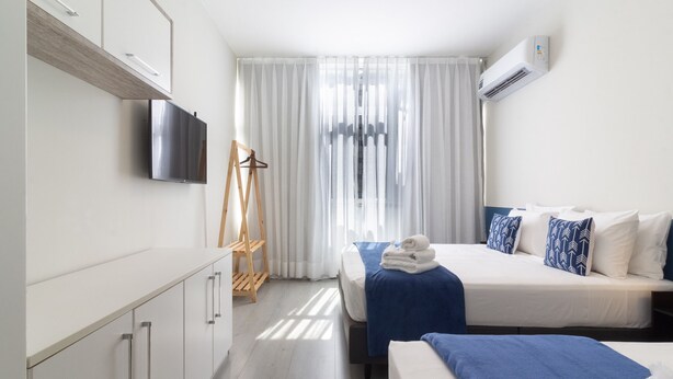 Une chambre d’hôtel avec un lit, un téléviseur fixé au mur, une chaise en bois et une fenêtre avec des rideaux légers.