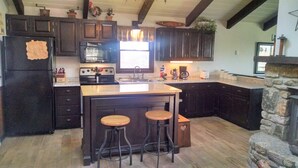A kitchen with dark wooden cabinets, a central island with stools, a stone fireplace, and a large window providing natural light. - CR-22 Harbeson Haven Peaceful 4 BR Chalet-Style Home Very well maintained (Beech Mountain)