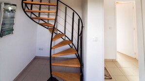 Una escalera de caracol con escalones de madera y barandillas de metal negro en una habitación con paredes blancas y suelo de baldosas.