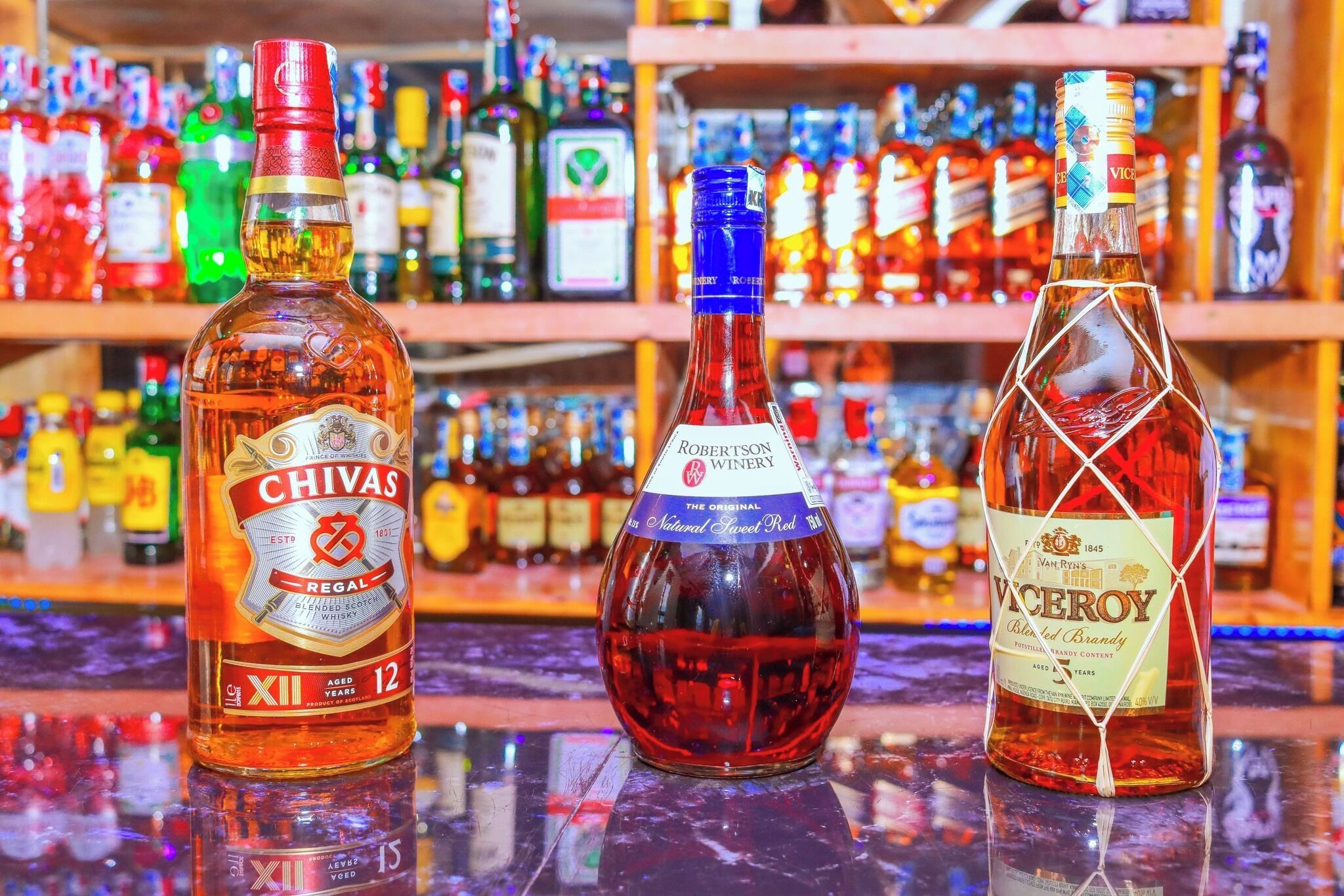 Three bottles of whiskey on a bar counter with shelves of various liquor bottles in the background.