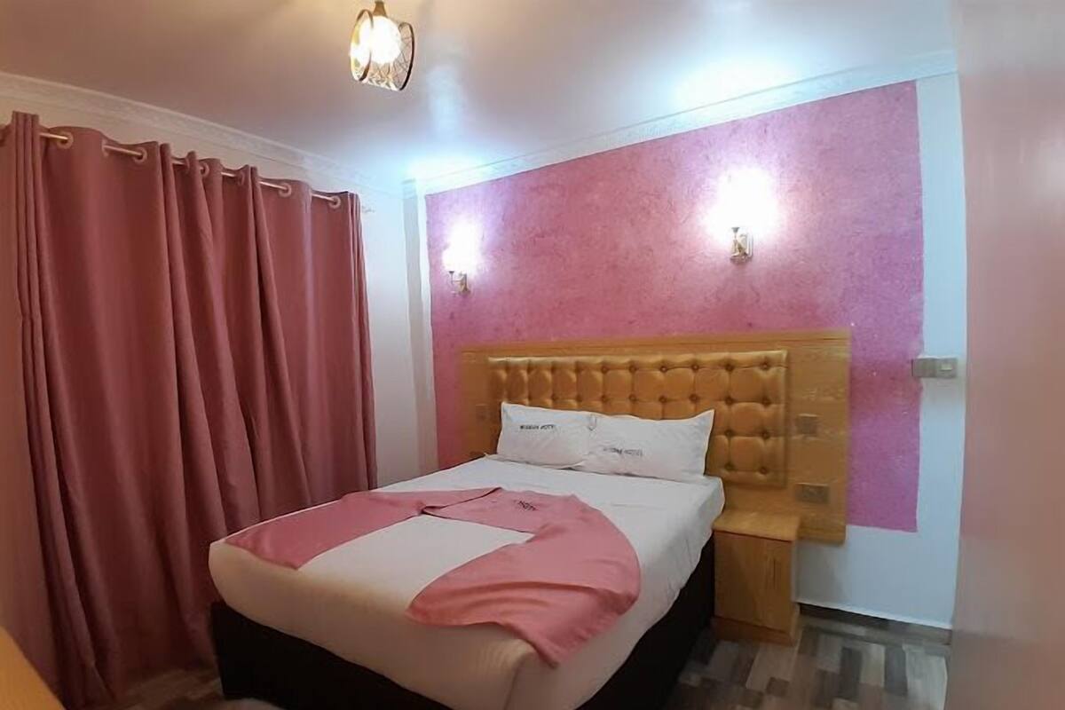 Quarto com cama, cortinas cor-de-rosa, cabeceira de madeira e mesa de cabeceira.