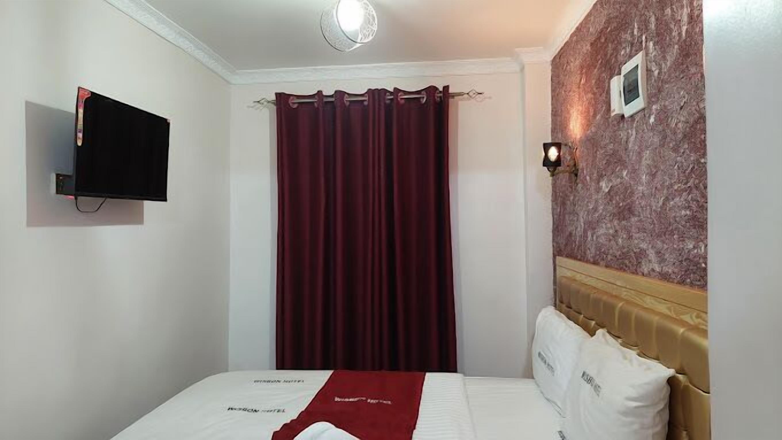Una habitación de hotel con cama, televisor montado en la pared, cortina roja y pared con diseño de mármol.