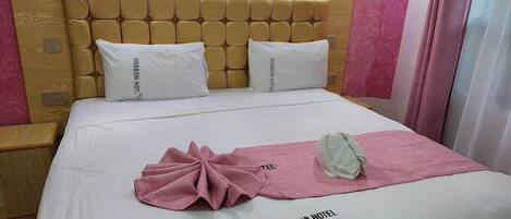 A hotel room with a wooden headboard, two white pillows, a pink towel, and a white blanket with "WISDOM HOTEL" text.