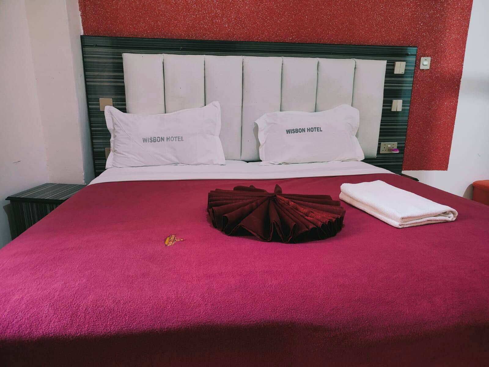 A hotel room with a bed, two pillows with "WISBON HOTEL" text, a red blanket, and a folded towel.