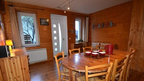Interior de una cabaña de madera con una mesa redonda, sillas de madera y una pequeña cocina.