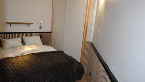 A bedroom with a bed, pillows, a blanket, a wooden headboard, a wall-mounted shelf, and a radiator.