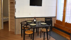 A dining area with a table set for four, chairs, a television mounted on the wall, and a bed in the adjacent room.