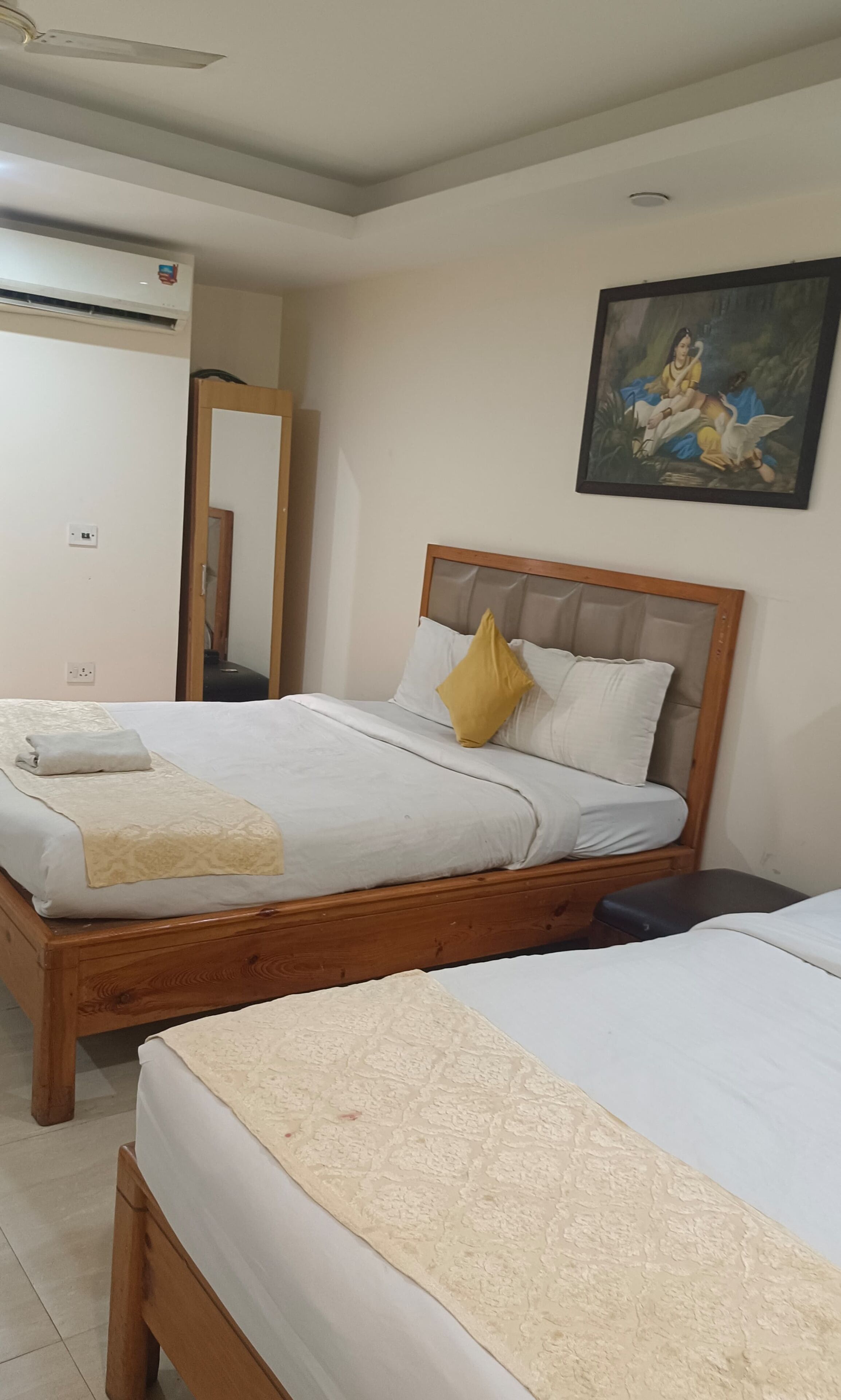 Een hotelkamer met twee houten bedden, een plafondventilator, een airconditioningapparaat, een spiegel en een schilderij aan de muur.