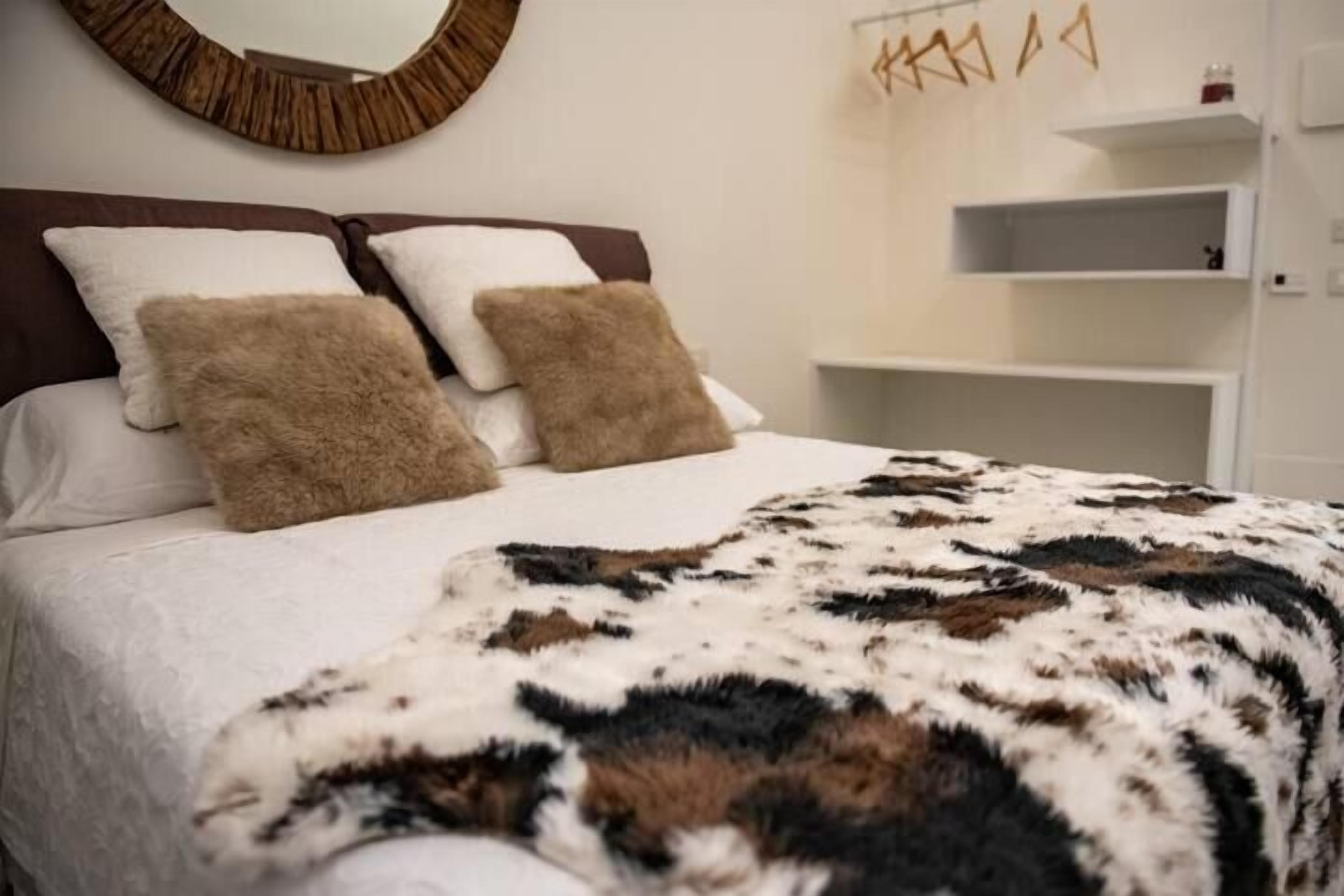 Un lit avec du linge de lit blanc, deux oreillers marron et une couverture à motifs. On y voit une tête de lit en bois, un miroir rond accroché au mur et une étagère blanche sur laquelle repose un petit objet.