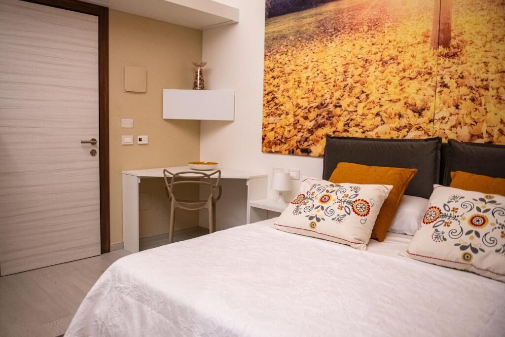A hotel room with a bed, a desk, and a chair. There is a large painting on the wall and a door to the left.