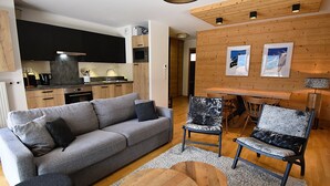 Una sala de estar moderna con un sofá gris, dos sillones estampados, una mesa de centro de madera y una cocina al fondo.