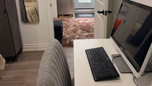 Ett modernt rum med ett vitt skrivbord, en dator och en stol. På väggen hänger en spegel, det finns en dörr som leder till ett annat rum och ett fönster med persienner.