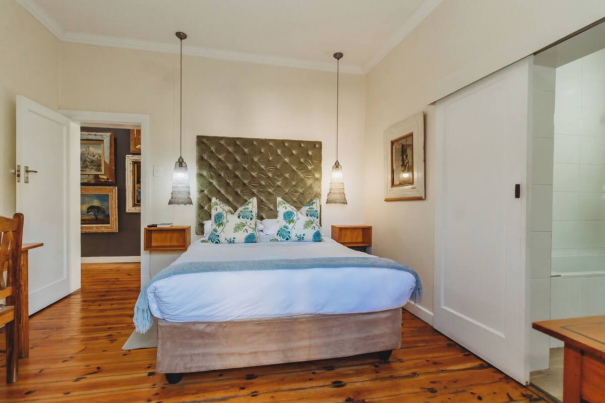 A bedroom with a large bed, wooden flooring, a tufted headboard, bedside lamps, and a door leading to another room.