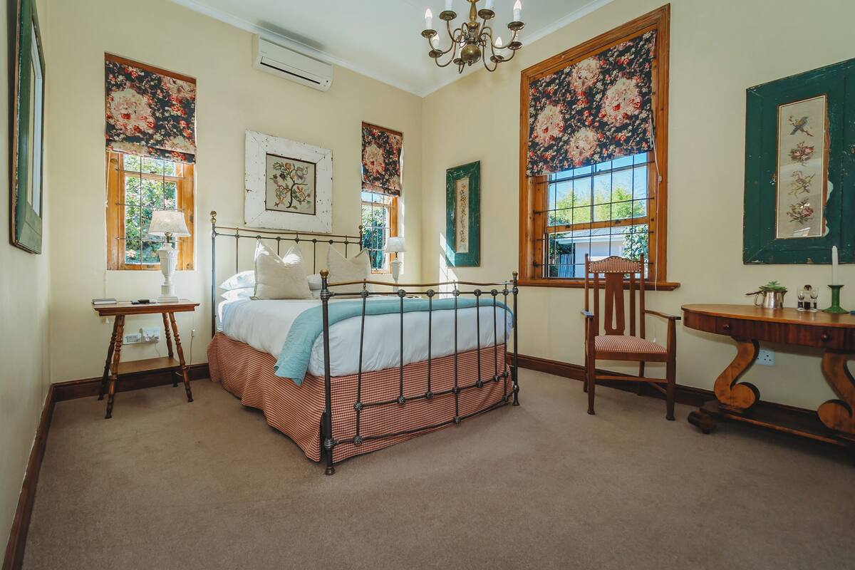 A bedroom with a bed, a chair, a table, and large floral windows.