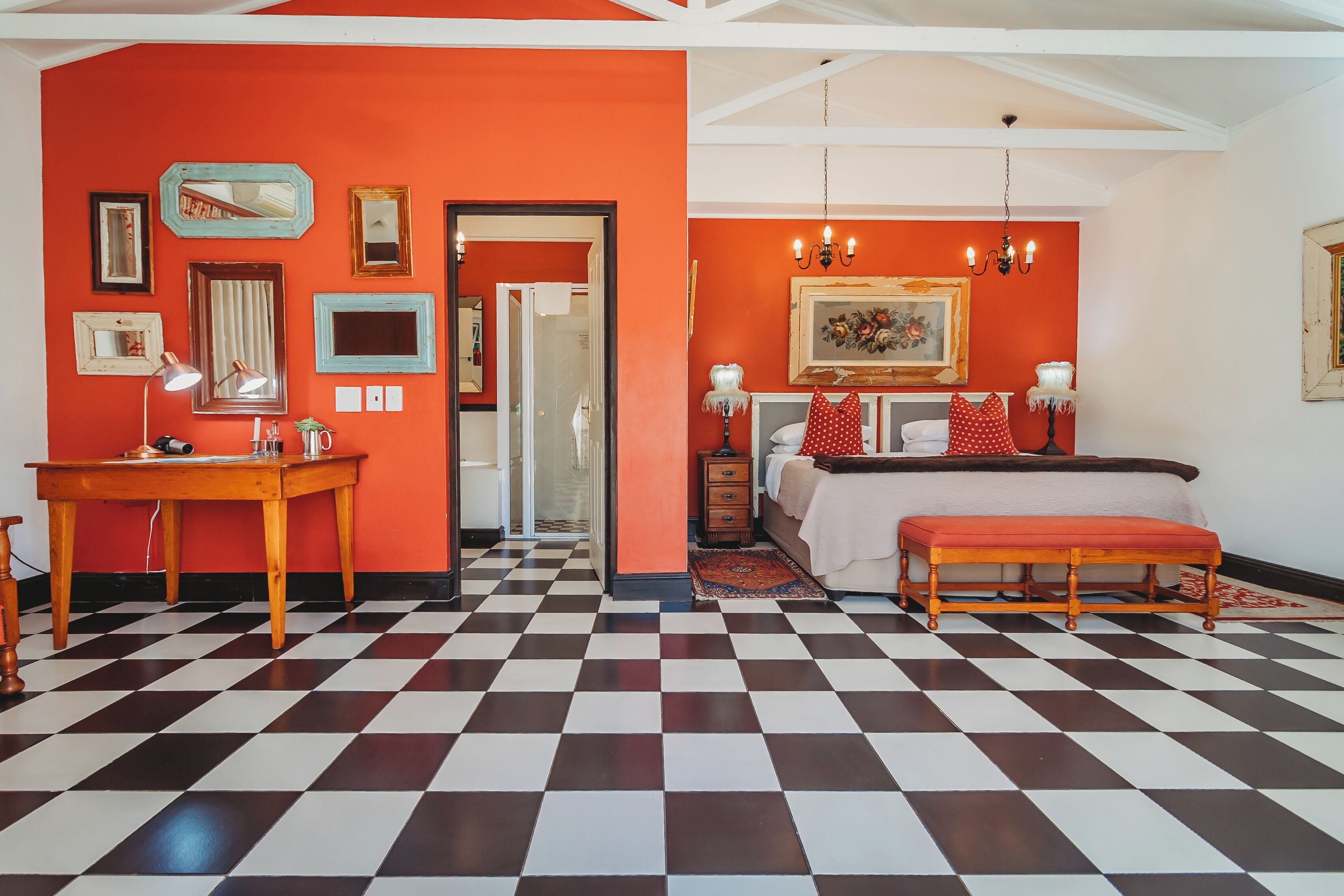 A room with a checkered floor, a bed with red and white bedding, a wooden bench, and a desk with a lamp.