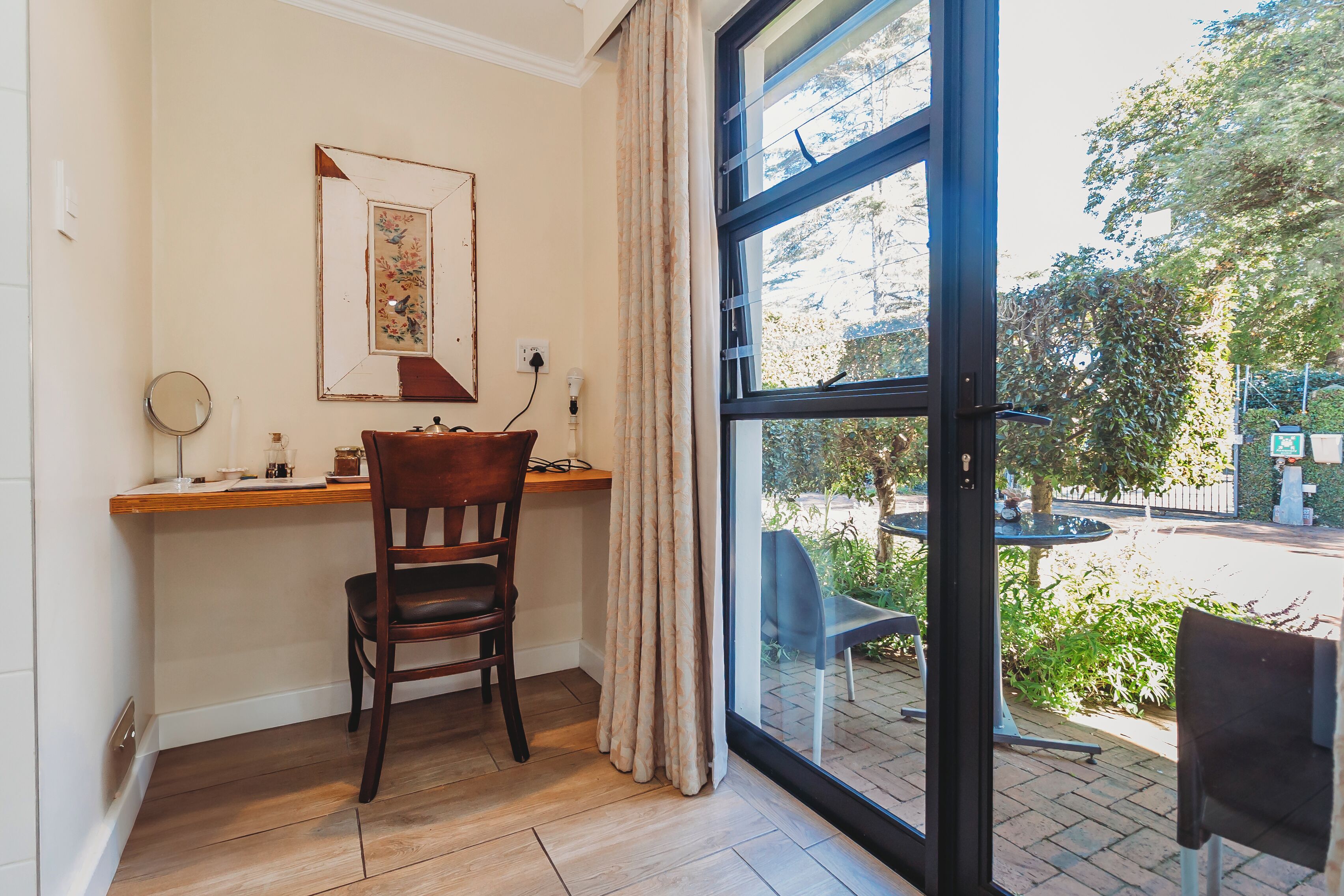 A room with a wooden desk, a chair, a framed picture, and a large window with a view of an outdoor seating area.