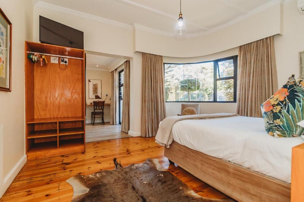 A bedroom with a wooden floor, a bed with a floral pillow, a wardrobe, and a view of the outdoors through a window.