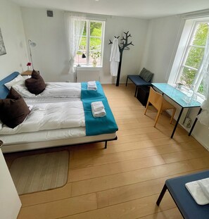 A hotel room with two beds, a small table, a bench, and a window with a view of greenery.