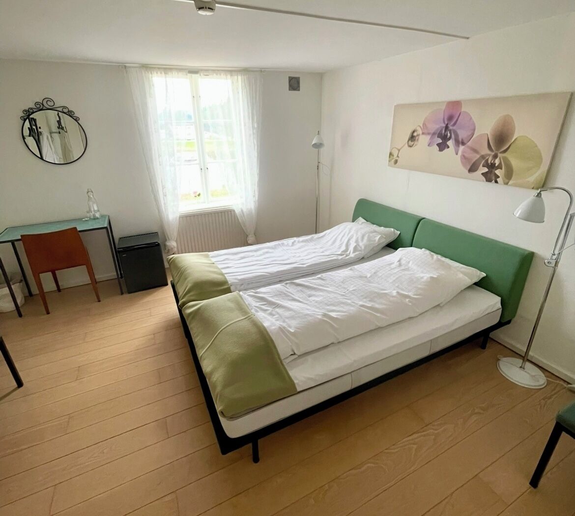 Habitación con una cama individual, cabecera verde, un escritorio con silla, un radiador pequeño y una ventana con cortinas traslúcidas.