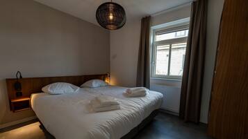 Una habitación de hotel con cama, mesita de noche, lámpara y ventana con cortinas.