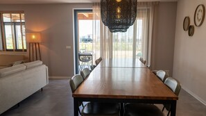 Een eetgedeelte met een lange houten tafel, zes stoelen, een bank, een lamp en uitzicht door een glazen deur.