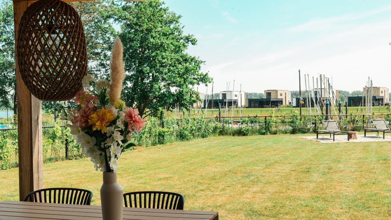An outdoor patio with a wooden table and chairs, a wicker lamp, and a vase of flowers. In the background, there is a grassy area with a fence, trees, and buildings.