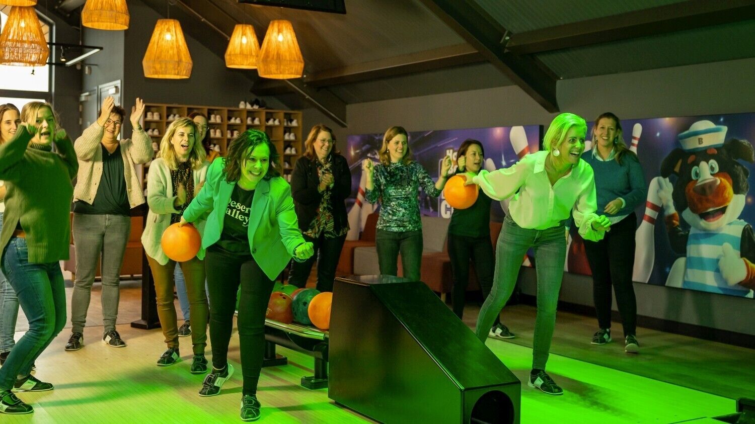 A bowling alley with multiple lanes, a green-lit floor, and a mascot in the background.