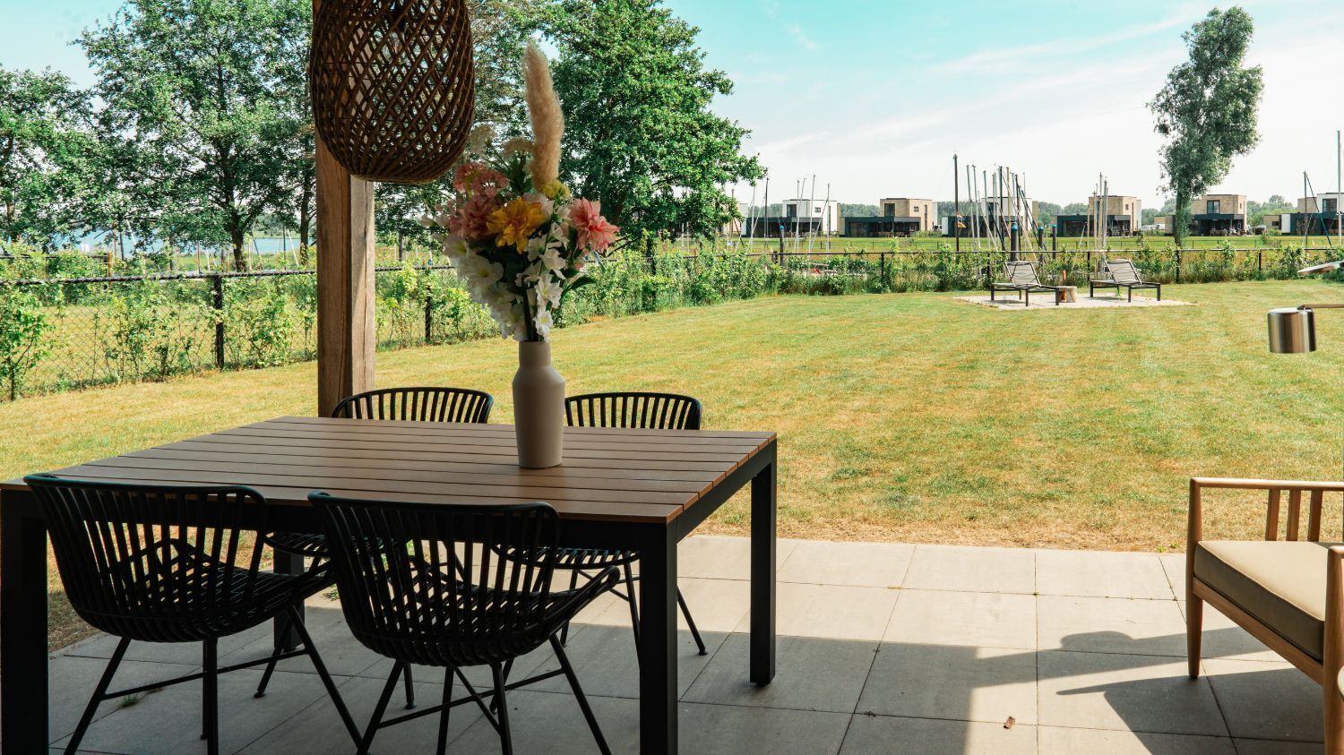 Une terrasse extérieure avec une table et des chaises en bois, un vase rempli de fleurs et une vue sur un espace verdoyant, avec des arbres et des bâtiments en arrière-plan.