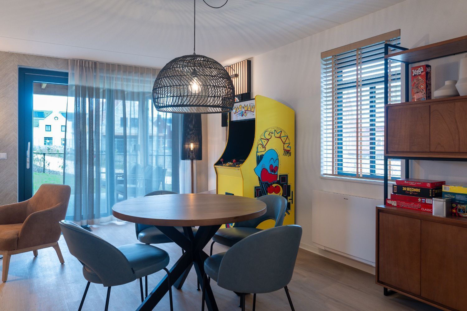 A modern living room with a round wooden table, four grey chairs, a black hanging light fixture, a yellow cabinet with a cartoon character, and a window with blinds.