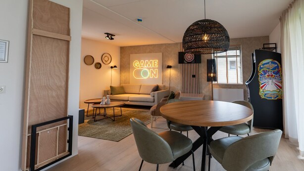 Una sala moderna con una mesa redonda de madera, sillas tapizadas en gris, un sofá y un letrero de neón que dice "GAME ON".