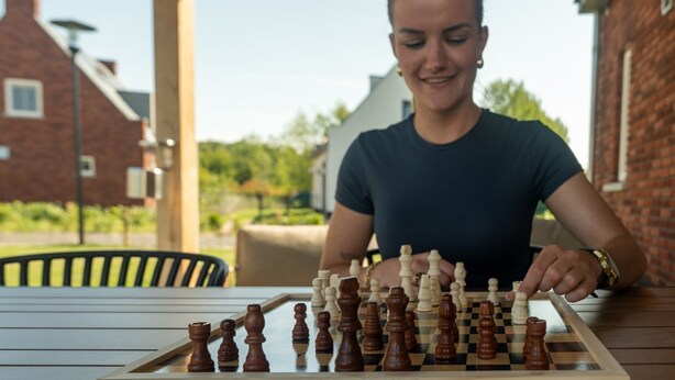 Iemand speelt schaak aan een buitentafel, met een helder uitzicht op een bakstenen huis en groen in de achtergrond.