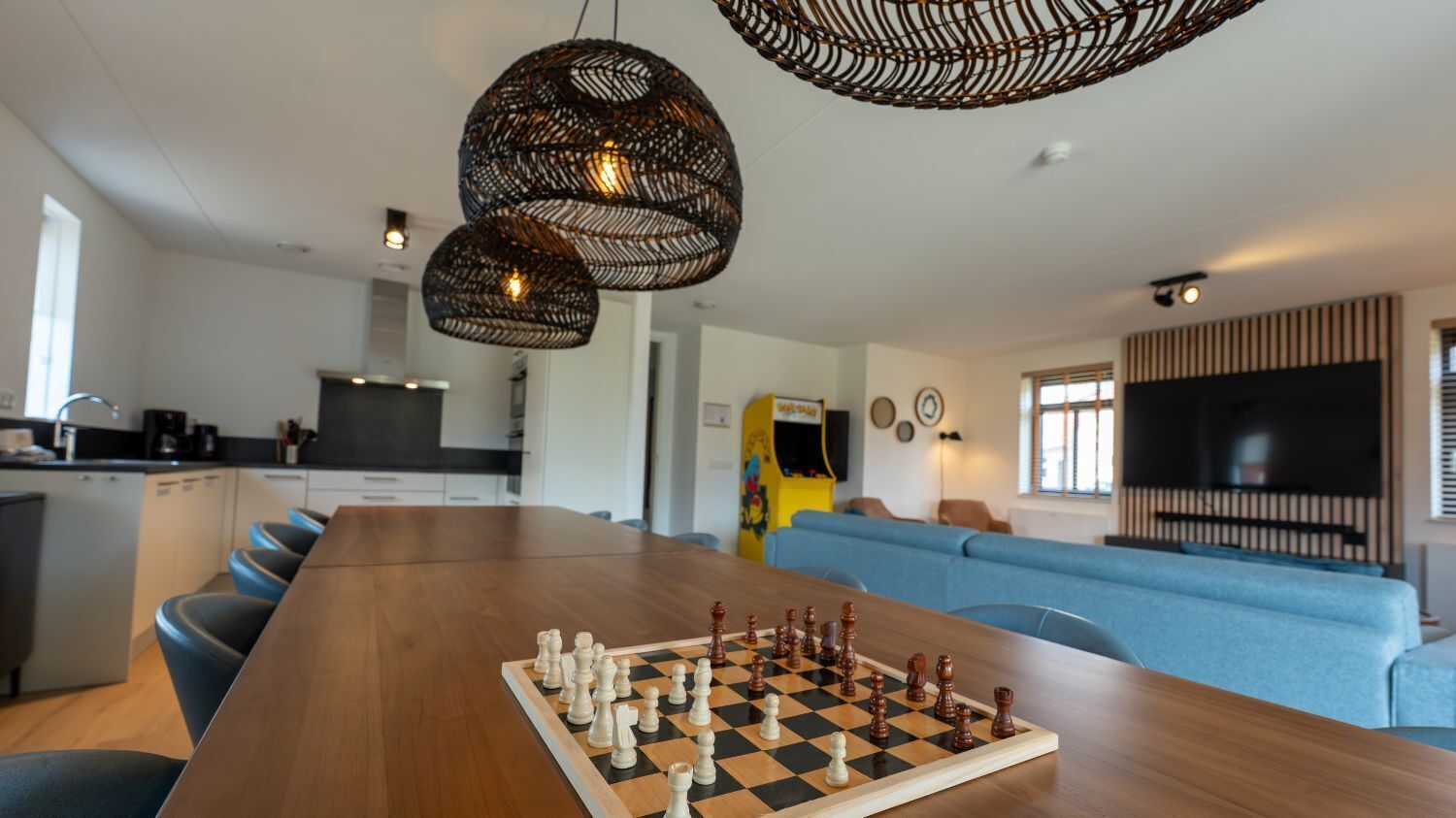 A modern living space with a wooden dining table set for a game of chess, a kitchen area, and a seating area with a large flat-screen TV.