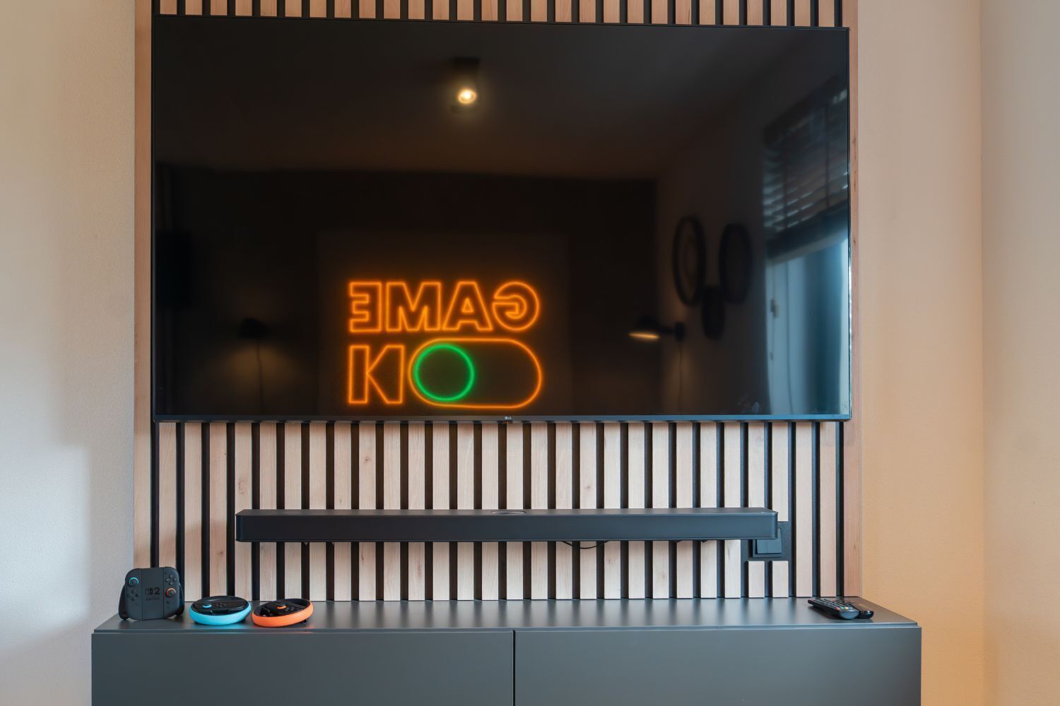 Une pièce moderne avec un néon mural indiquant « GAME KO  », un téléviseur fixé au-dessus d’une étagère en lattes de bois et une console de jeux posée sur le sol.