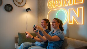 Twee personen spelen een videospel op een bank, met een neonbord waarop 'GAME ON' staat in de achtergrond.
