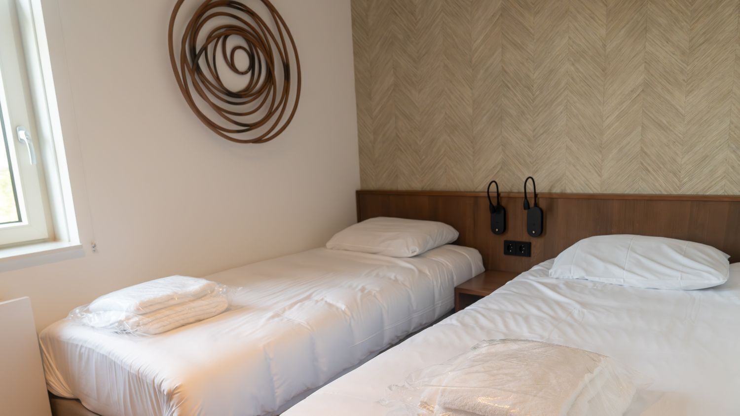 A hotel room with two single beds, a wooden headboard, a wall-mounted phone, and a window with blinds.