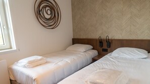 Une chambre d’hôtel avec deux lits simples, une tête de lit en bois, un téléphone fixé au mur et une fenêtre avec des stores.
