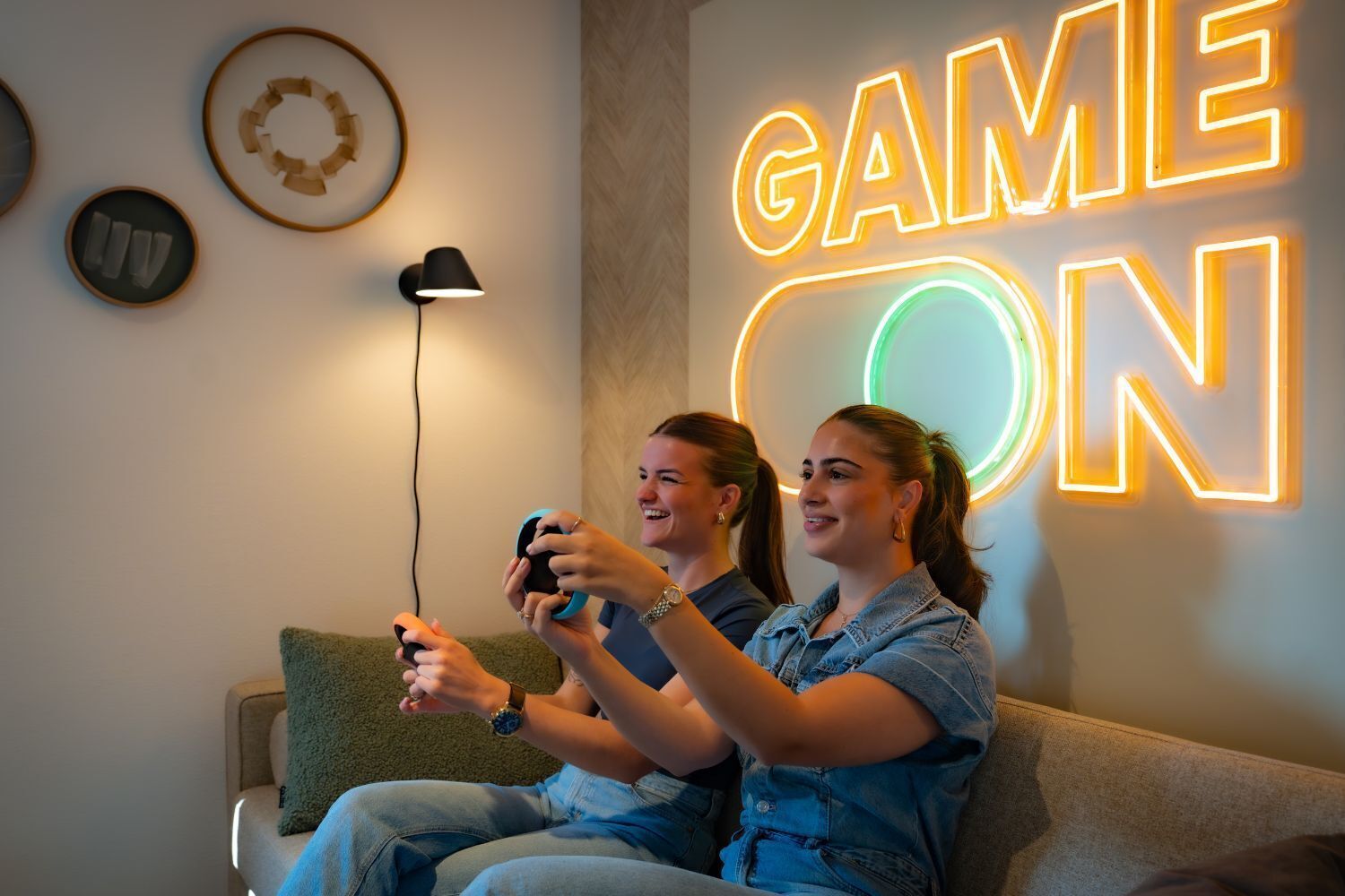 Two people playing video games on a couch with a neon sign that reads "GAME ON" in the background.