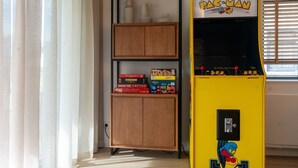 Una habitación con una máquina arcade Pac-Man, un mueble de madera con juegos de mesa y una ventana con persianas.