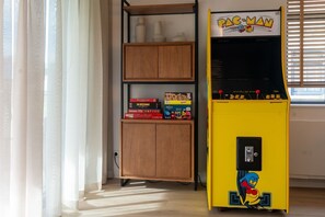 A room with a Pac-Man arcade machine, a wooden cabinet with board games, and a window with blinds.