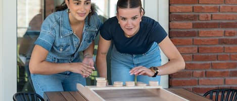 Zwei Personen spielen ein Brettspiel an einem Holztisch, im Hintergrund ist eine Backsteinmauer zu sehen.