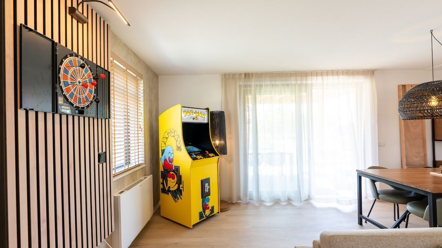 Een kamer met een dartbord, een Pac-Man spelautomaat, een eettafel en een raam met gordijnen.
