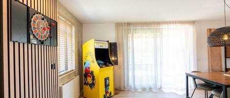 Een kamer met een dartbord, een Pac-Man spelautomaat, een eettafel en een raam met gordijnen.