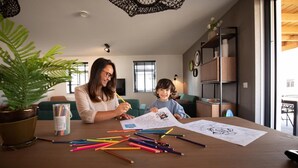 Una mujer y un niño coloreando en una mesa con varios lápices de colores, rodeados de plantas en macetas y un interior moderno.