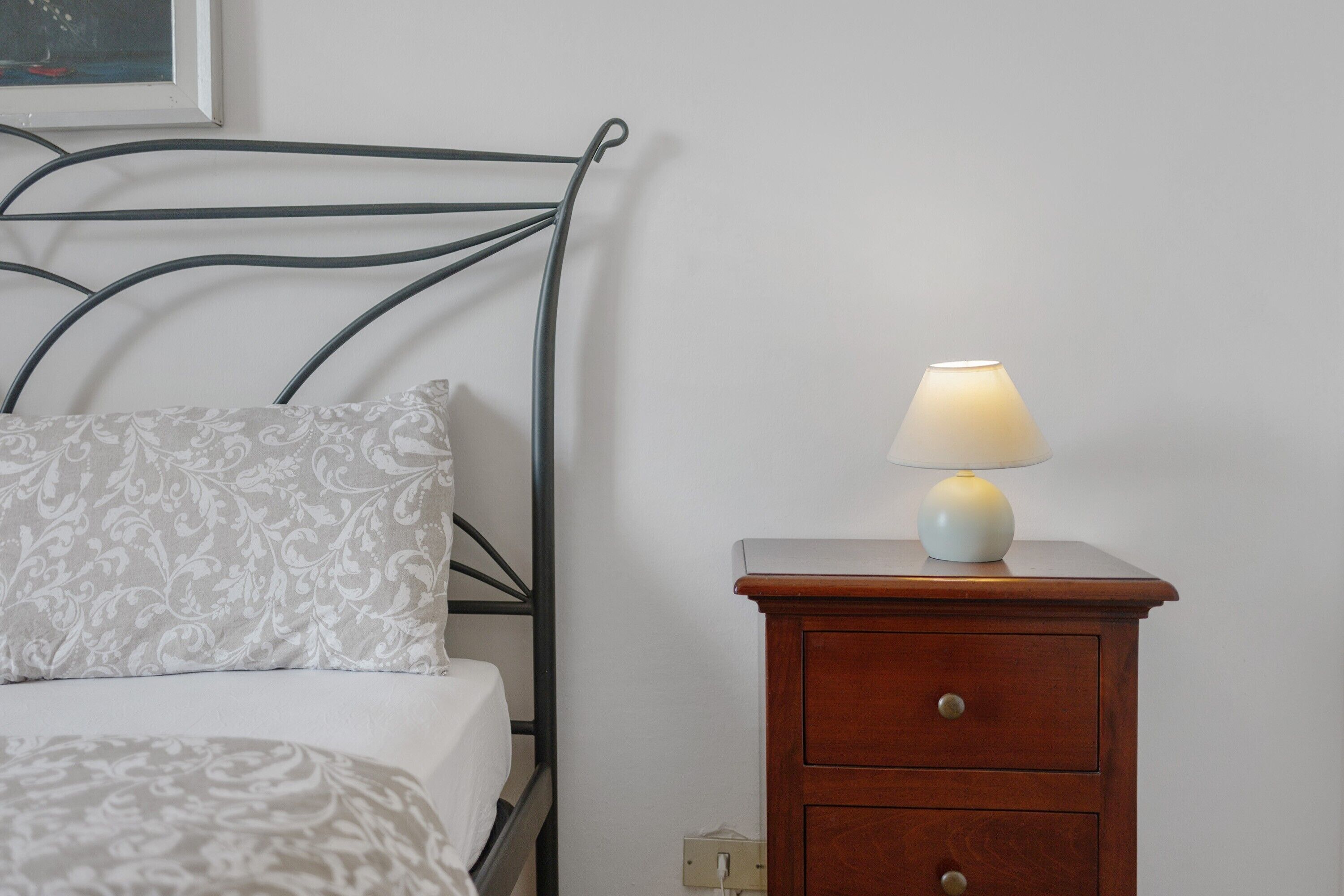 A bed with a patterned pillow, a bedside table with a lamp, and a wall-mounted light switch.