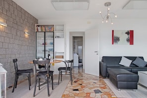 A modern living room with a sofa, a dining table with chairs, a bookshelf, and a patterned floor. - Holiday Home 'Belvedere36' with Lake View, Garage, E-Bikes & Air Conditioning (Bedero)
