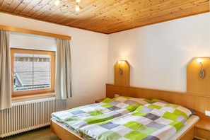 A bedroom with a wooden ceiling, a window with curtains, a bed with a checkered bedspread, and two wall-mounted light fixtures.