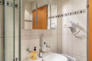 Un baño con cabina de ducha de vidrio, un lavamanos con dispensador de jabón, una toalla y un secador de pelo en un soporte fijado a la pared.