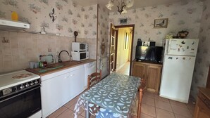 Een kleine keuken met een fornuis, magnetron, spoelbak en koelkast. Een eettafel met stoelen staat in het midden.