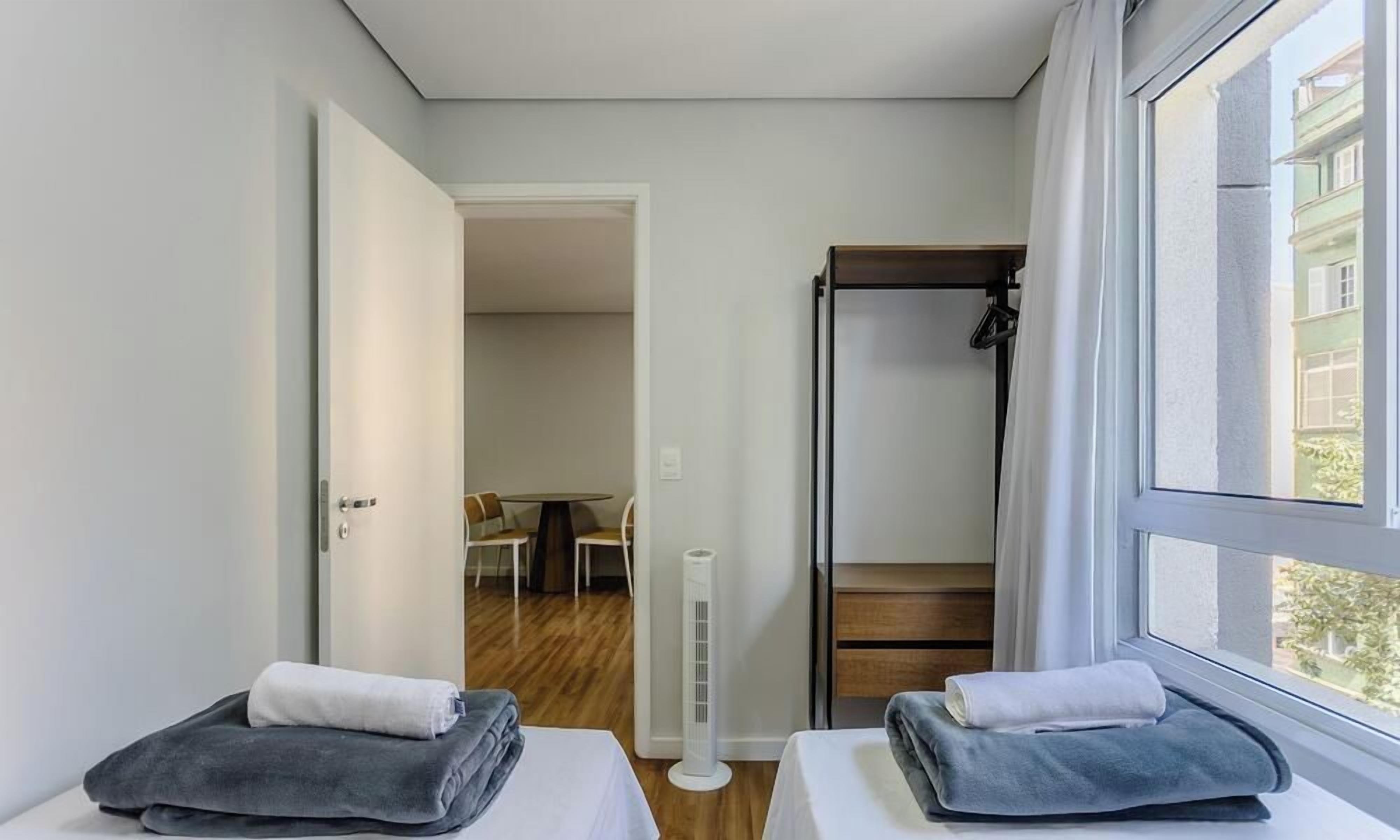 A hotel room with two beds, a standing heater, a window with a view, and a door leading to a dining area.