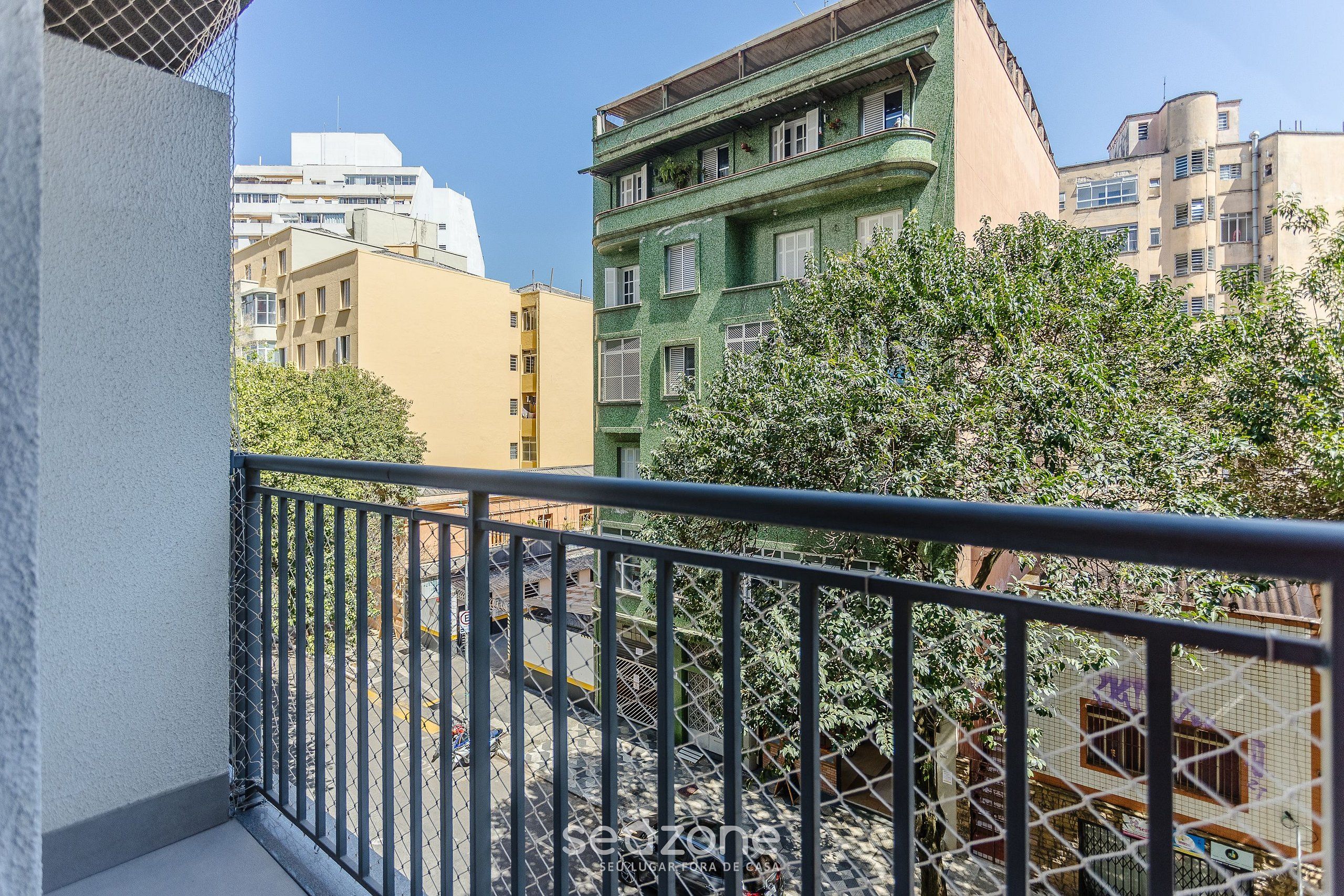 A balcony with a metal railing overlooking a street with buildings and trees.